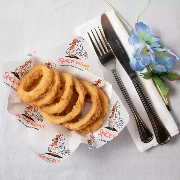 Golden onion rings in a Spice Wing branded paper basket, with cutlery and a blue flower.