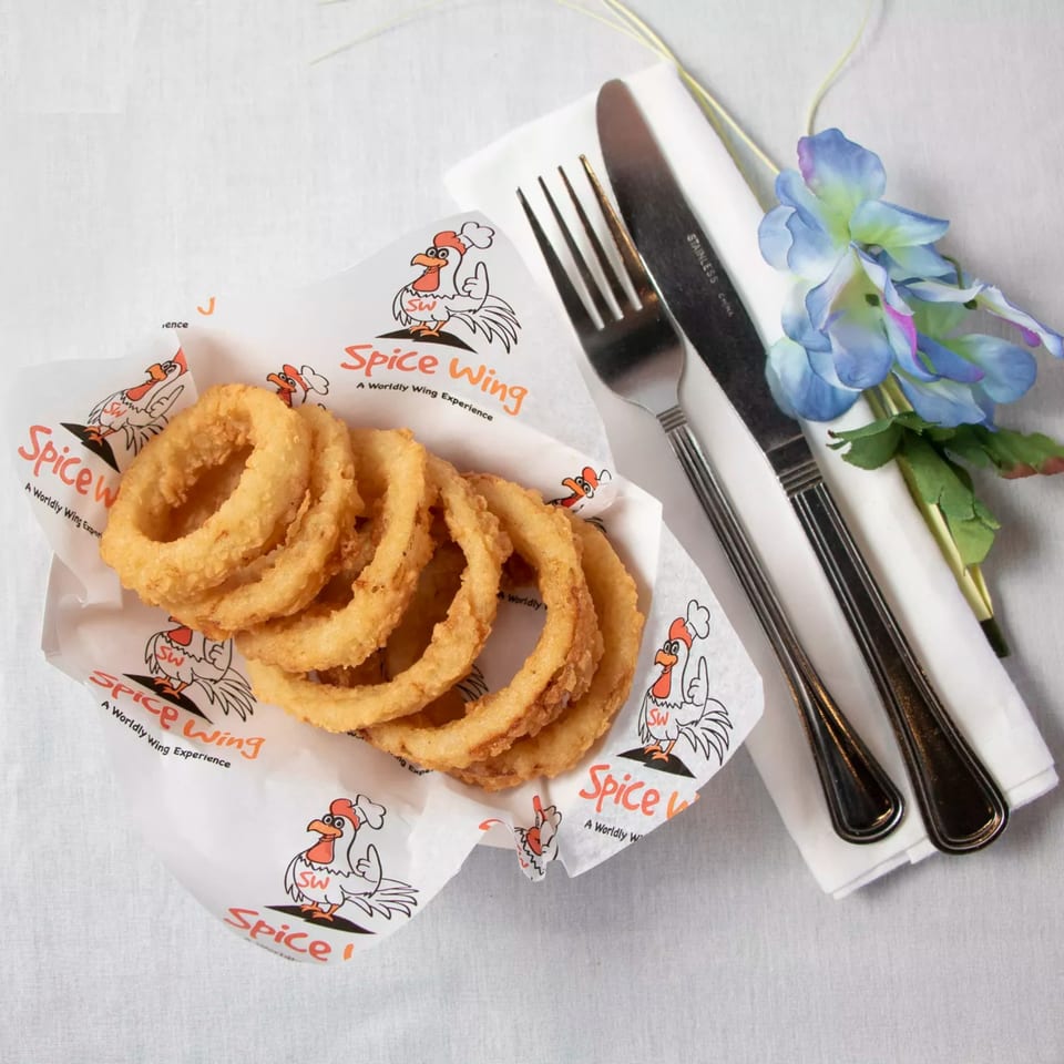 Golden onion rings in a Spice Wing branded paper basket, with cutlery and a blue flower.