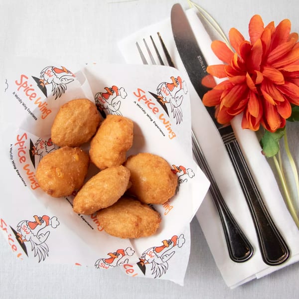 Six golden-brown Spice Wing boneless wings in a branded paper basket next to silverware and an orange flower.