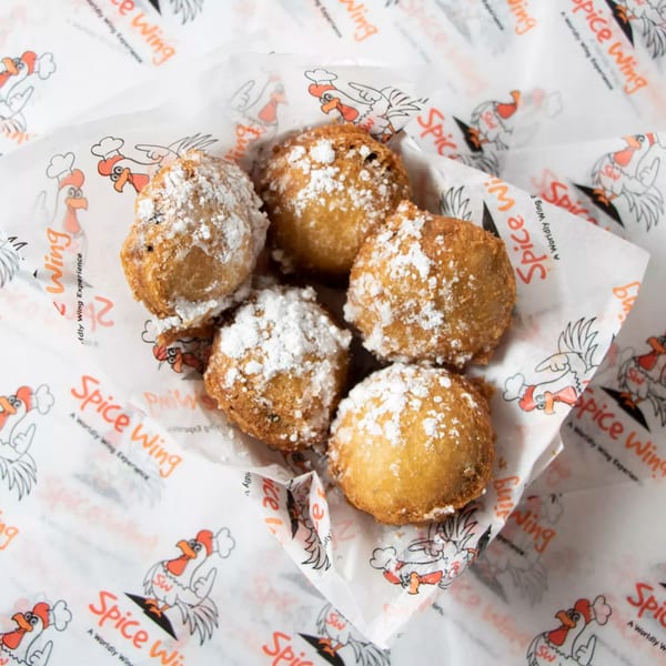 Five golden-brown hushpuppies, dusted with powdered sugar, in a basket lined with Spice Wing paper.