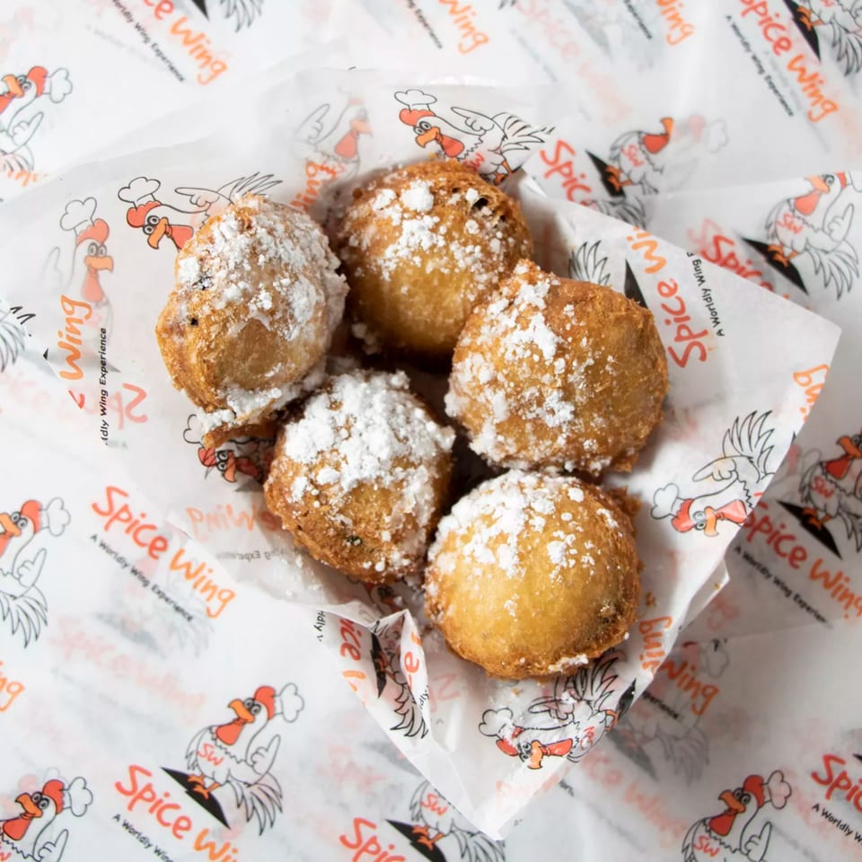 Five golden-brown fried dough balls, dusted with powdered sugar, in a Spice Wing branded paper container.